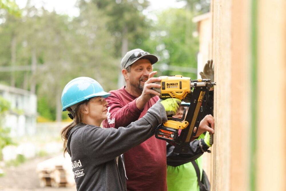 someone helping another person use a yellow nail gun on the wall of a house that is being built