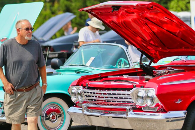 A teal and blue car parked with their hoods open and a man in a gray shirt and sunglasses walking by them