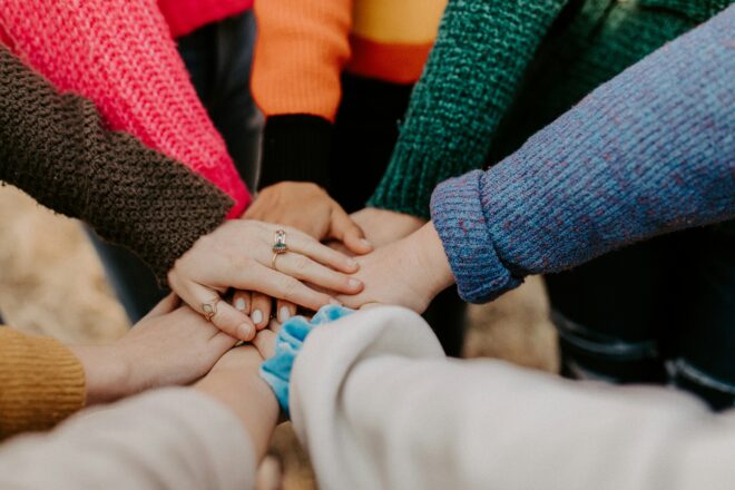 Arms in a circle with multicolored sleeves putting their hands together in the center, hand on top has multiple gold rings