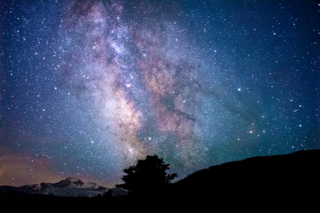 The outline of a hill with a large tree in front of a large night sky filled with orange, dark purple, and blue galaxies and stars