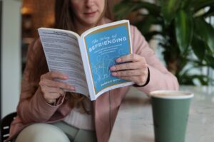 Woman in pink jacket sitting in a coffee shop reading a blue book with a green coffee cup in front of her.