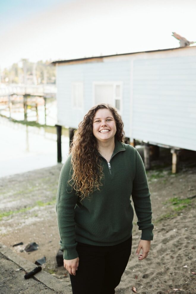 Person with long brown hair in a dark green half zip sweater in front of a body of water and a light blue house on stilts.