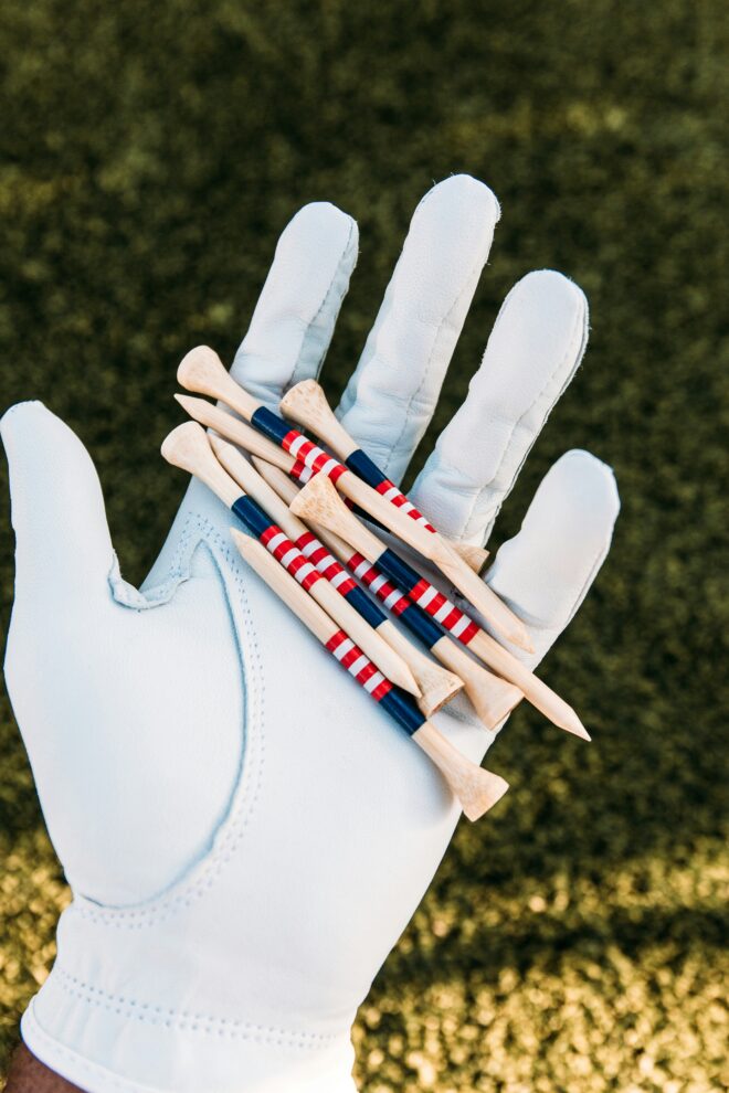 Hand in bright white glove atop a dark green grass background holding tan golf tees with red, white, and blue stripes.