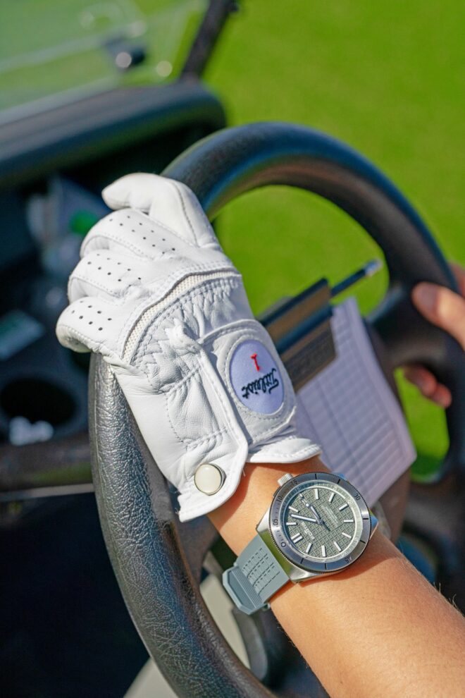 Hand in white glove with gray watch on wrist holding black steering wheel of a golf cart that you can see blurred out in the background with bright green grass.