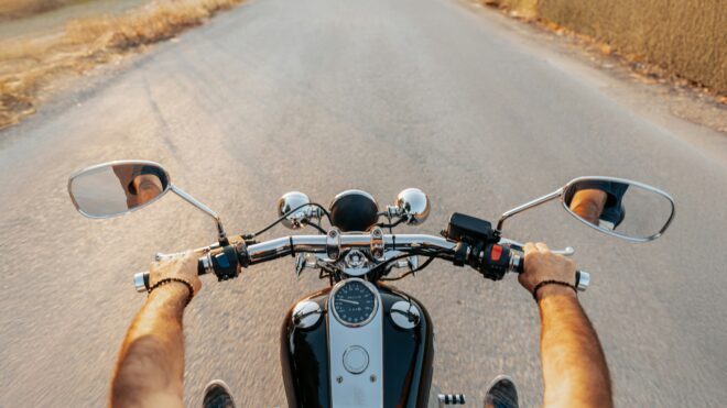 First person point of view, two arms holding onto the bars of a black and silver motorcycle as it drives on a gray paved road surrounding by yellow grass.