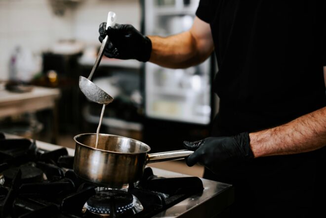 Person in black shirt with black latex gloves on pours white sauce from silver ladle into silver sauce pot.